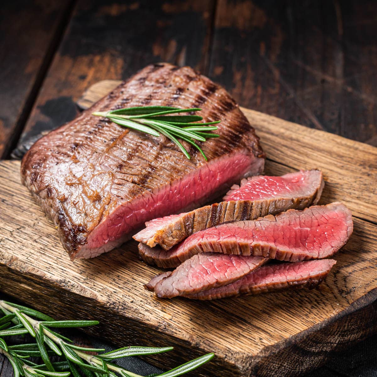 Grilled medium rare flank steak on a rustic wooden cutting board with a sprig of rosemary.