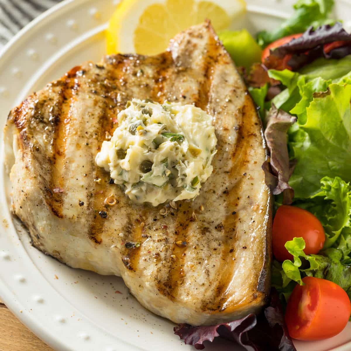 Grilled swordfish on a plate with a green salad and a lemon slice.